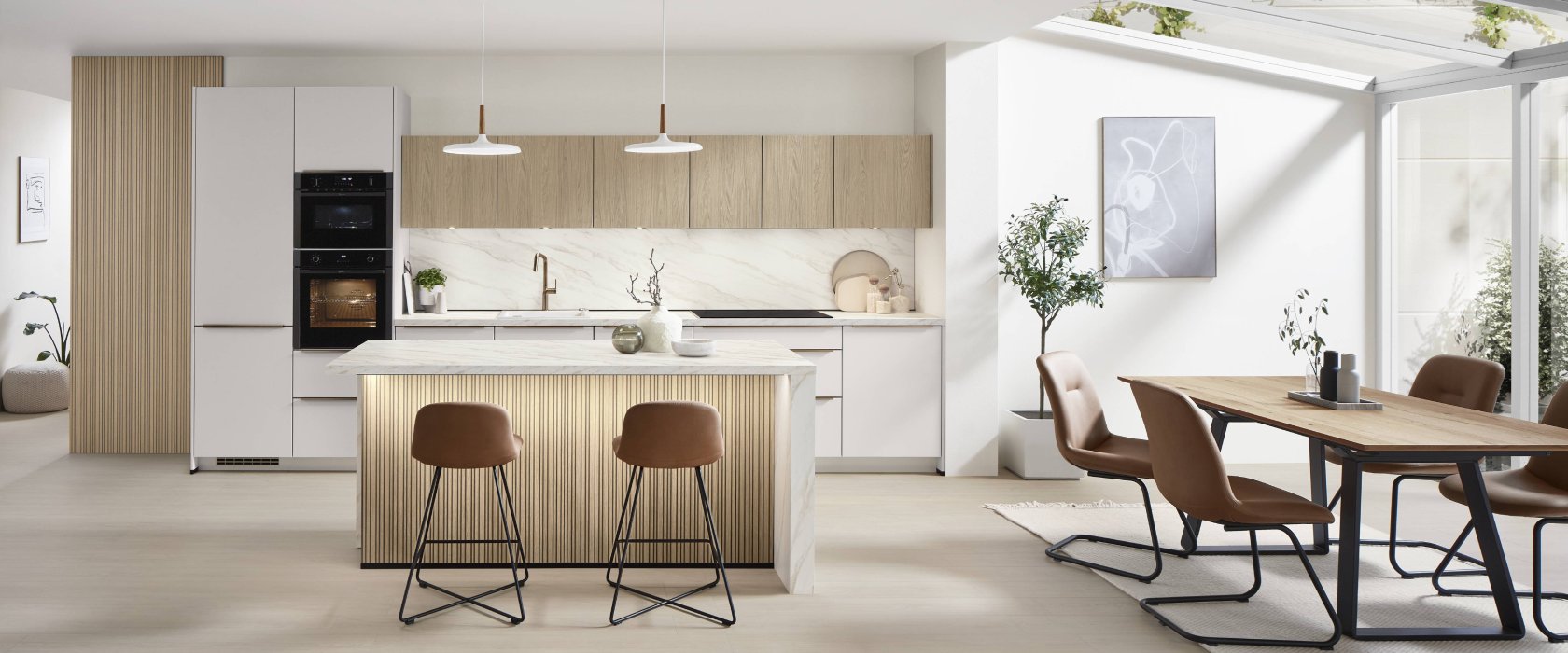Velký celkový pohled na designovou kuchyni Nobilia Softline 509 v provedení bílá Perfect matná s mramorovou deskou Carrara Bronze a navazujícím jídelním stolem.