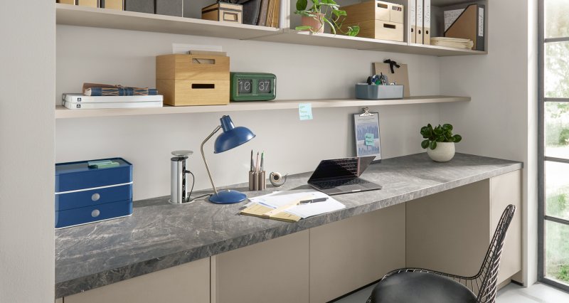 Detailní pohled na moderní domácí pracovnu integrovanou do výklenku, která kombinuje béžové spodní skříňky, tmavě šedou strukturovanou pracovní desku a otevřené nástěnné police.