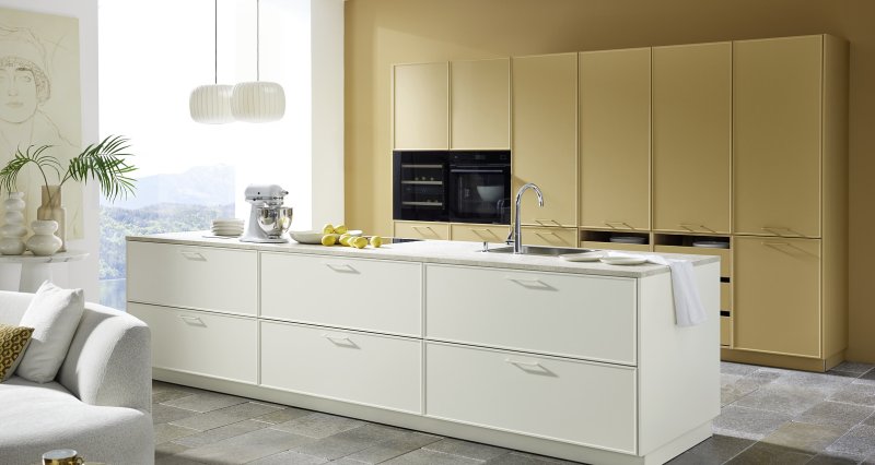 Prostorná kuchyně Nolte Evora kombinující velký ostrůvek v krémovém laku Magnolia softmatt a vysokou stěnu v odstínu Caramel softmatt.