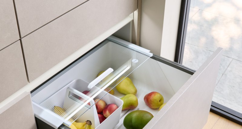 Otevřená kuchyňská zásuvka z laminátu Terra taupe vybavená bílým plastovým systémem Pantry-Box s průhledným krytem, plná čerstvého ovoce.