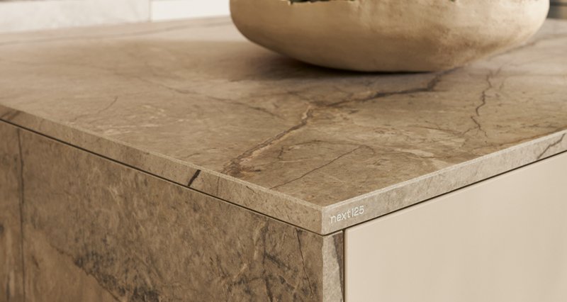 Detailní záběr na rohový spoj a strukturu luxusní kuchyňské pracovní desky s výraznou kresbou přírodního kamene a minimalistickým logem značky.