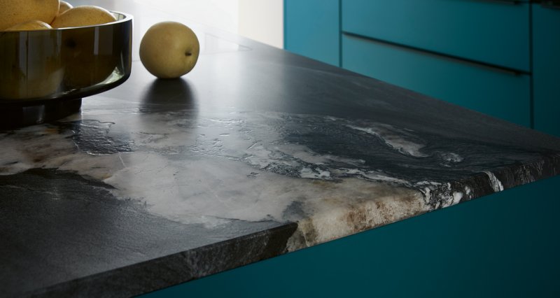 Detailní pohled na strukturu prémiové tmavé pracovní desky Systemo v dekoru Black Tempest s výraznou bílou a hnědou kresbou, ležící na petrolejově modrých skříňkách.