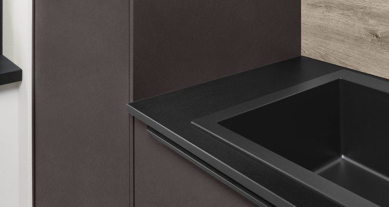 Detail rohu skříňky Nobilia Look v matném hnědém dekoru s černým úchopovým profilem a granitovým dřezem zapuštěným v tenké kompaktní desce