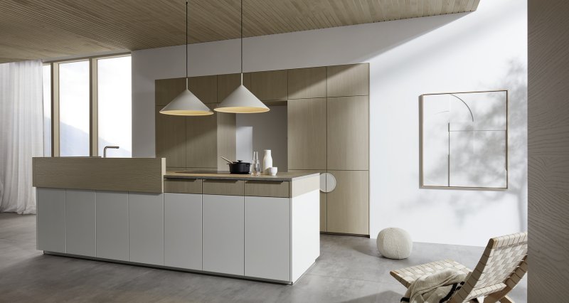 Širokoúhlá perspektiva kuchyně Nolte neo Leaf v kombinaci bílé matné barvy a jasanu pískového s designovým křeslem v popředí.