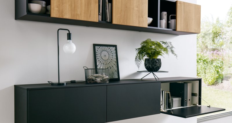 Černá matná závěsná TV skříňka s vyklápěcími dvířky, nad kterou je umístěna horní závěsná skříňka s designovými posuvnými čely v dekoru světlého dřeva.