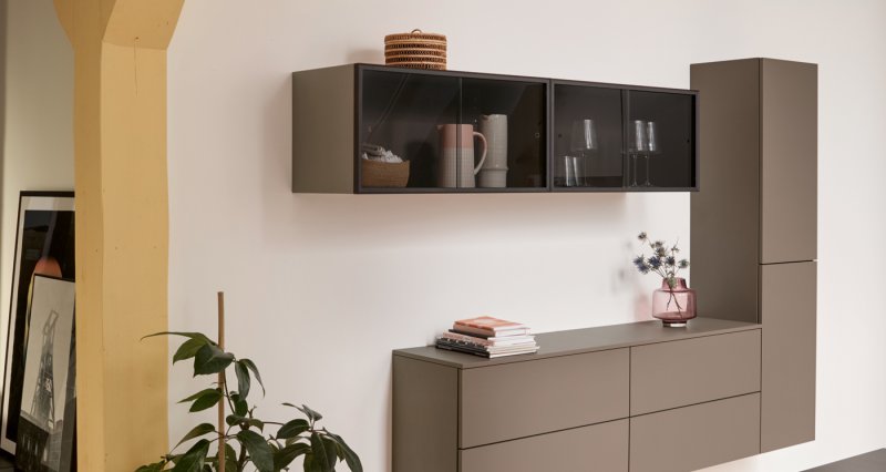 Moderní nástěnná sestava skříněk v matném šedohnědém laku s kouřovým sklem a plovoucí komodou u dřevěného sloupu