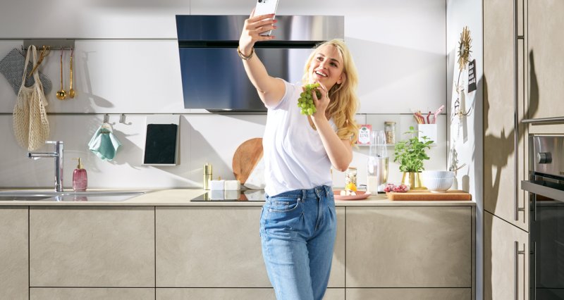 Mladá žena s hrozny vína fotící se na mobilní telefon v moderně zařízené kuchyni