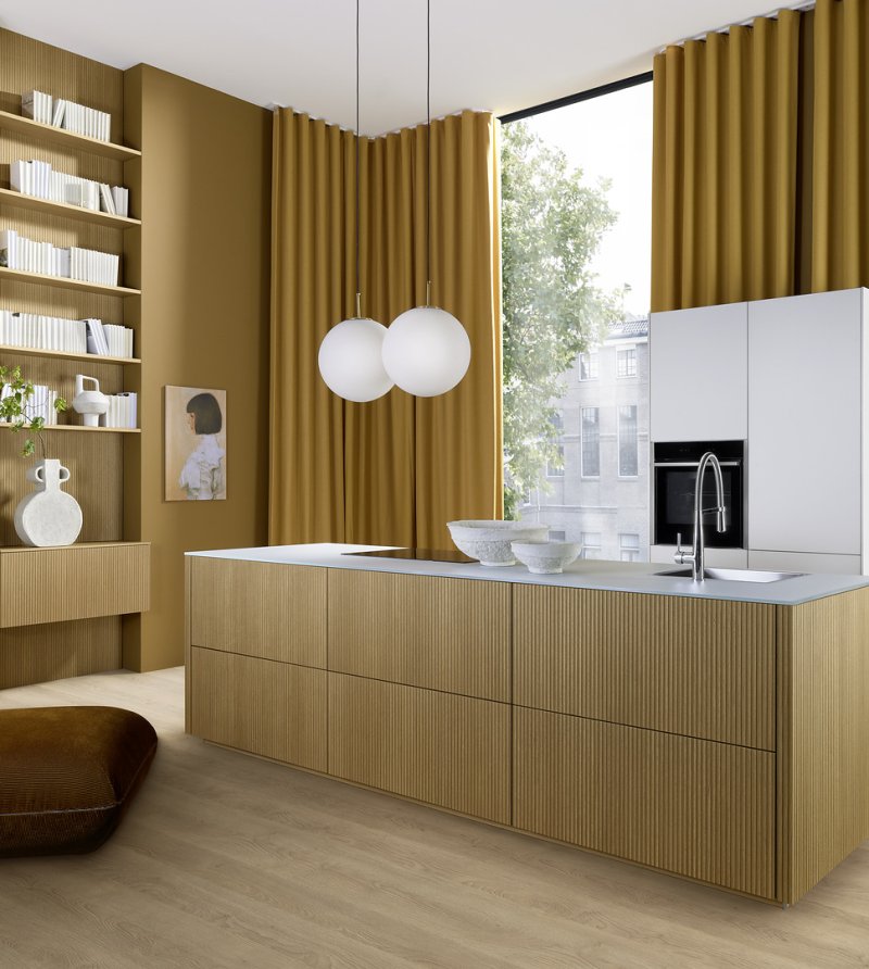 Prostorná kuchyně Nolte neo LODGE kombinující drážkovaný ostrůvek ze světlého dubu, pracovní desku Glas Tec Satin a vysoké bílé skříně.