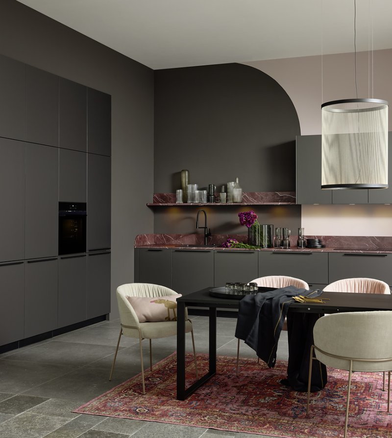 Přehledný pohled na designové uspořádání kuchyně Nolte Pearl s dominantní pracovní deskou z červeného mramoru a moderním jídelním stolem.