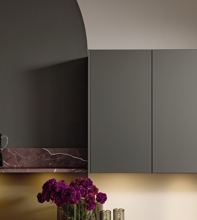 Detail minimalistických horních skříněk v odstínu Umbra metallic s integrovaným osvětlením nad pracovní deskou z červeného mramoru.