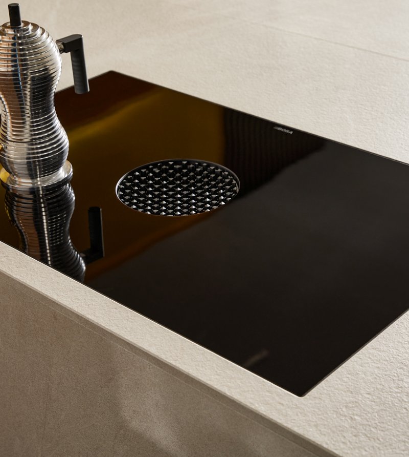 Detailní záběr na moderní varnou desku s integrovaným odvětráváním a designovým italským kávovarem na keramické pracovní desce Systemo.