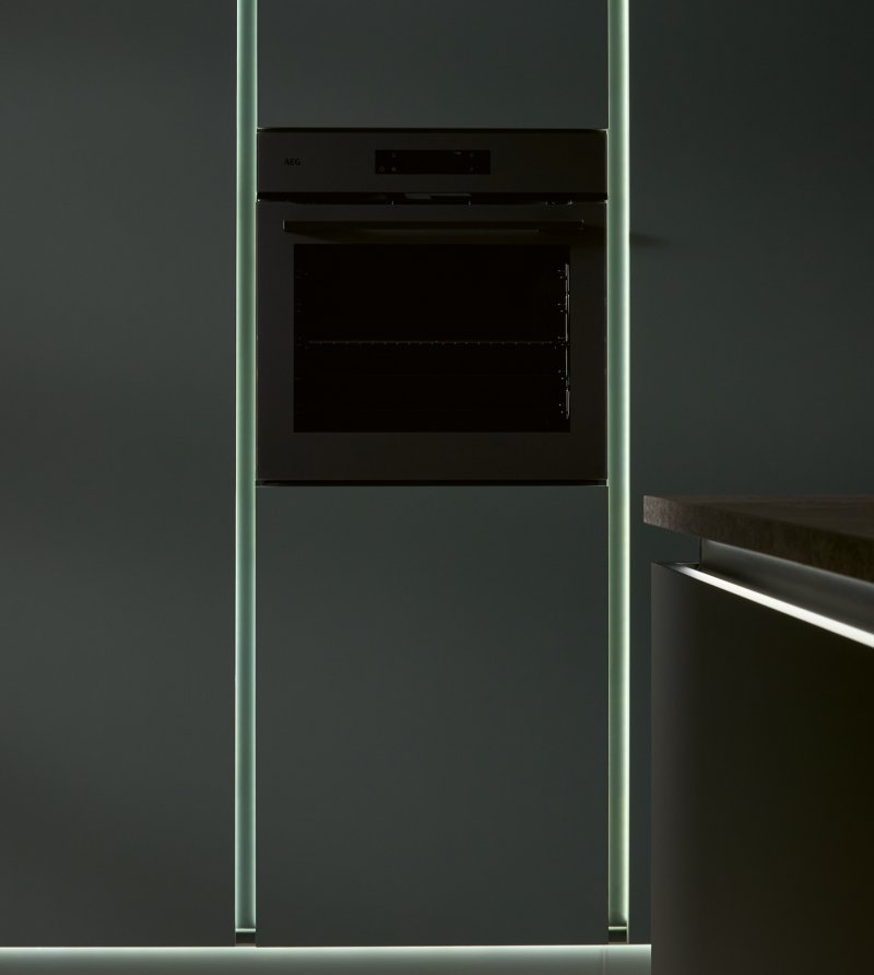 Detailní záběr na tmavou vestavnou troubu elegantně zasazenou do matné zelené stěny, podél které probíhají dva svislé svítící LED pruhy.
