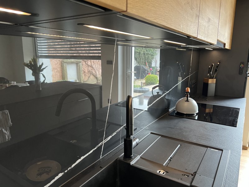 Detailní záběr na černou pracovní desku, nerezový dřez, minimalistickou černou baterii a vestavěné LED osvětlení pod horními skříňkami.