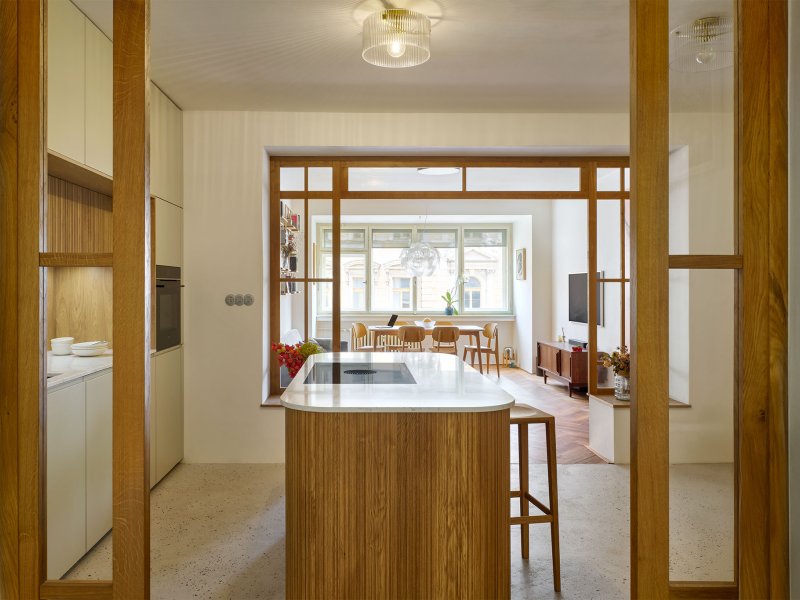 Pohled z obývacího pokoje skrz elegantní dřevěné prosklené dveře na moderní kuchyňskou linku s centrálním lamelovým ostrůvkem.