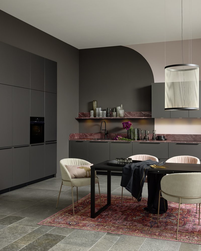 Přehledný pohled na designové uspořádání kuchyně Nolte Pearl s dominantní pracovní deskou z červeného mramoru a moderním jídelním stolem.