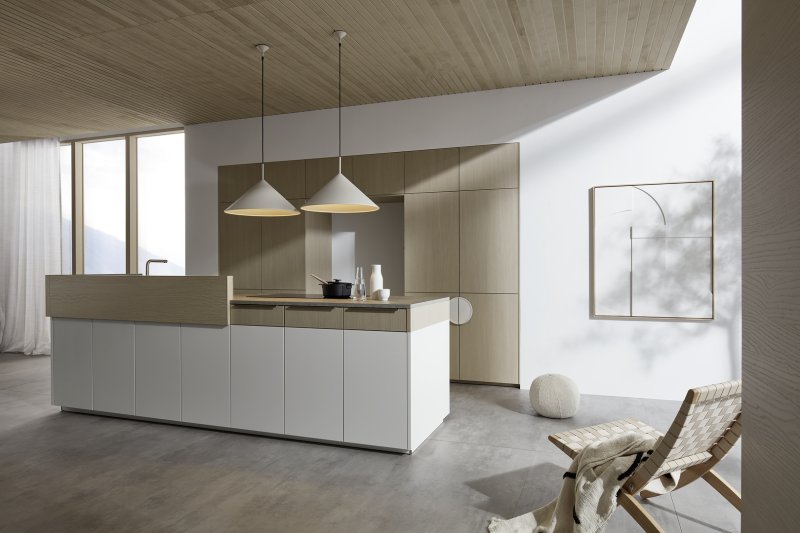 Širokoúhlá perspektiva kuchyně Nolte neo Leaf v kombinaci bílé matné barvy a jasanu pískového s designovým křeslem v popředí.