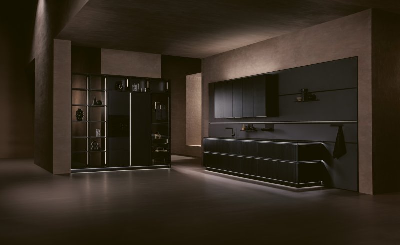 Řada spodních skříněk z dýhy dub onyx s rozsvícenými horizontálními úchytovými profily těsně pod tenkou skleněnou pracovní deskou.