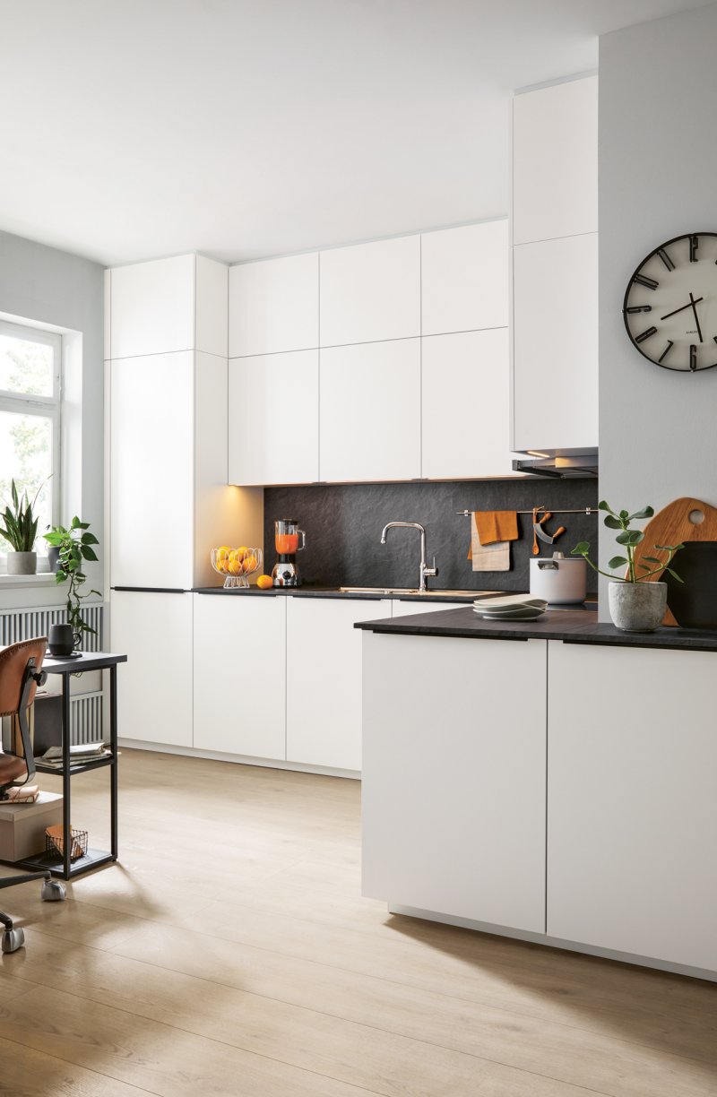 Pohled na moderní rohovou kuchyňskou linku v bílém matném designu s tmavou pracovní deskou, vestavnou troubou a hodinami na stěně