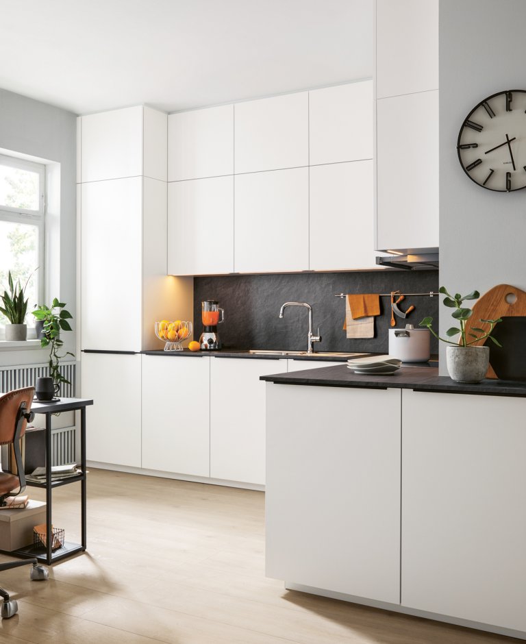 Pohled na moderní rohovou kuchyňskou linku v bílém matném designu s tmavou pracovní deskou, vestavnou troubou a hodinami na stěně