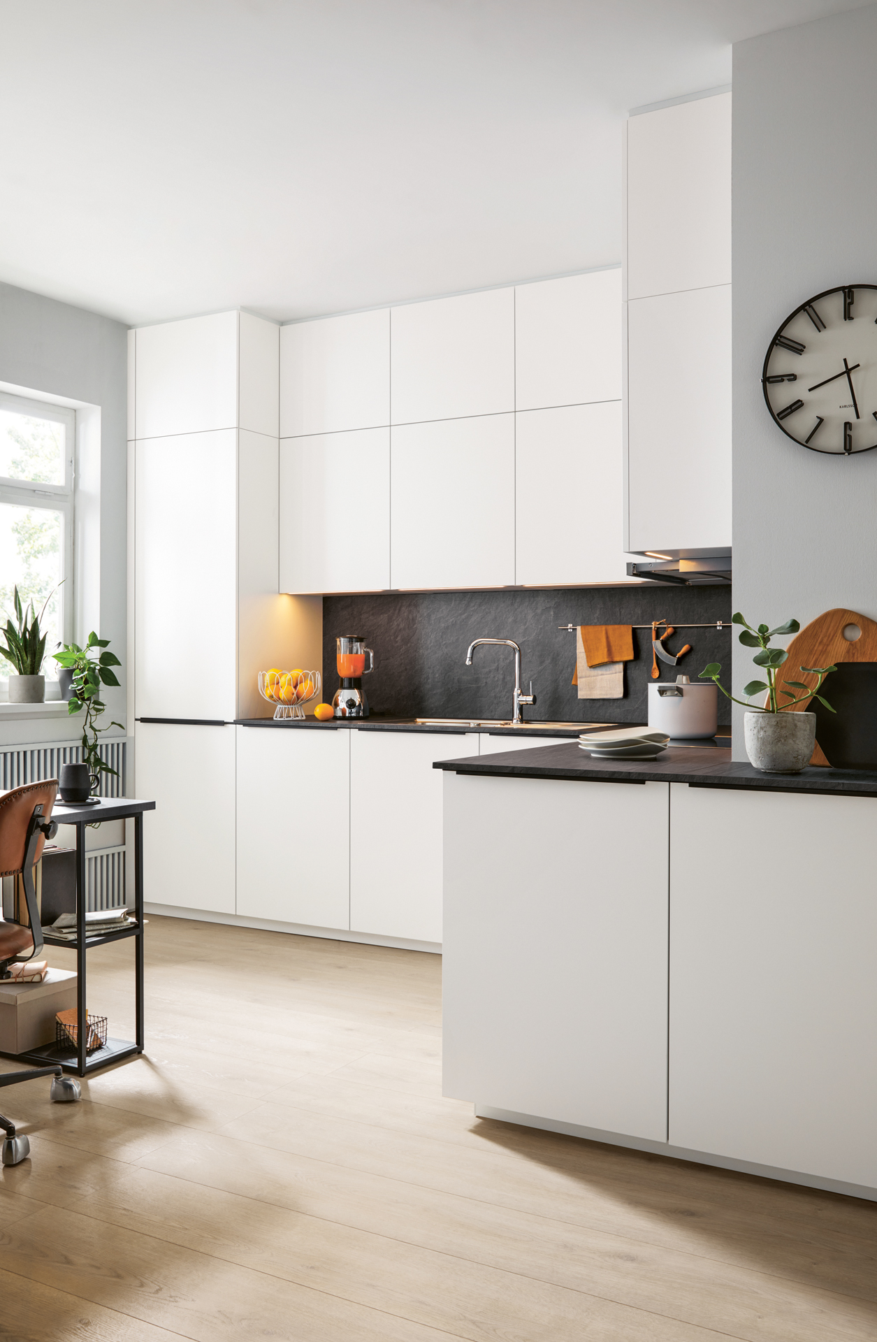 Pohled na moderní rohovou kuchyňskou linku v bílém matném designu s tmavou pracovní deskou, vestavnou troubou a hodinami na stěně